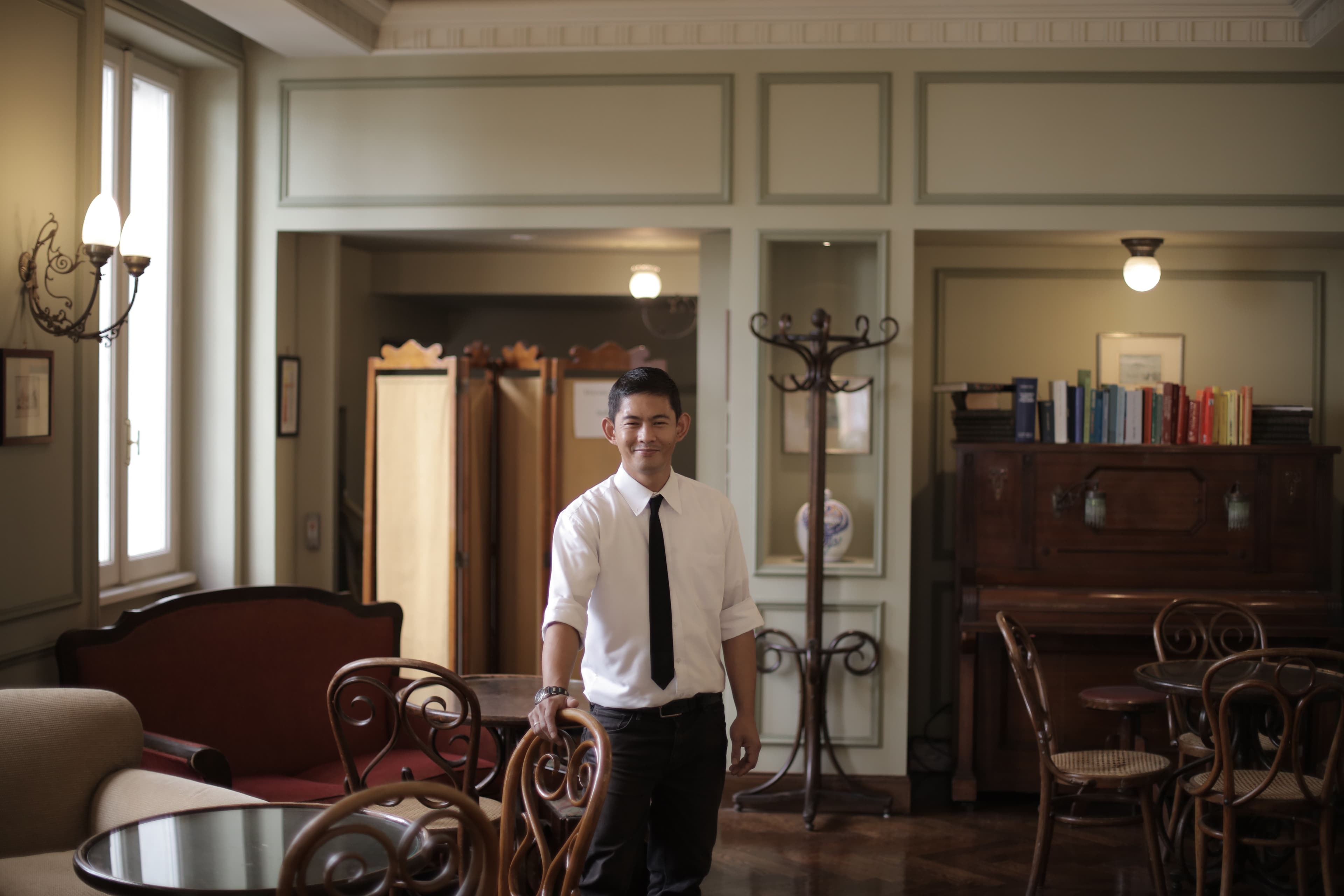 A relaxed restaurant host in a shirt and tie in a cozy dining room.